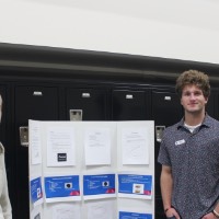 Two students smiling by a laptop and poster board.